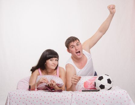 Young girl and a guy in bed. Beautiful girl has a manicure, man watches football on TV.