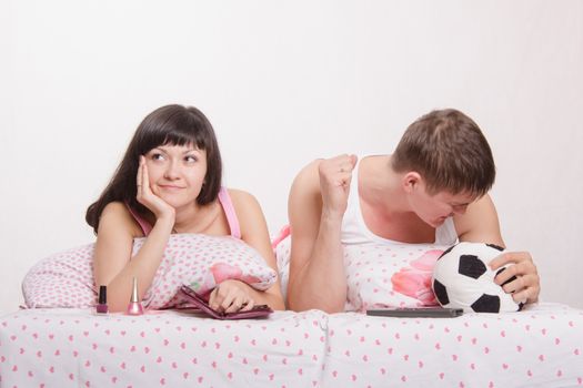 Young girl and a guy in bed. Beautiful girl has a manicure, man watches football on TV