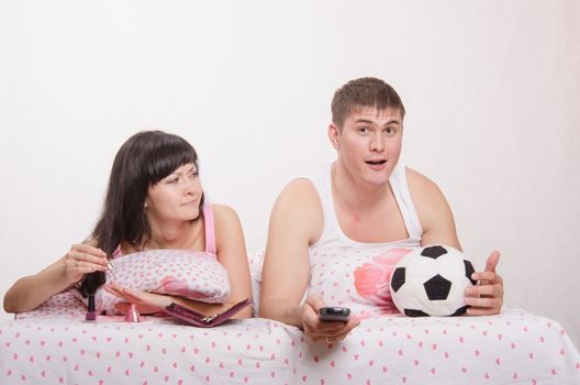 Young girl and a guy in bed. Beautiful girl has a manicure, man watching soccer on TV