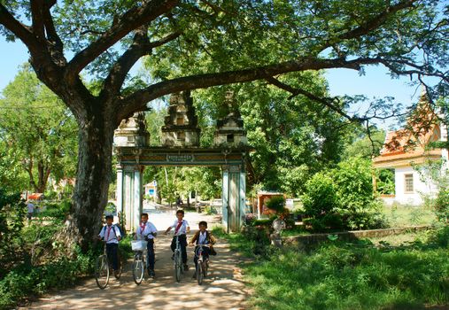 AN GIANG, VIET NAM- SEPT 20: Group of Asian primary pupil riding bike to Khmer village gate, ancient gate with large green tree, big brach make shade, kid happy with friendship, Vietnam, Sept 20, 2014