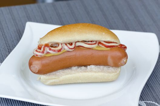 Hot dog with ketchup and mustard in bread roll on a white plate.