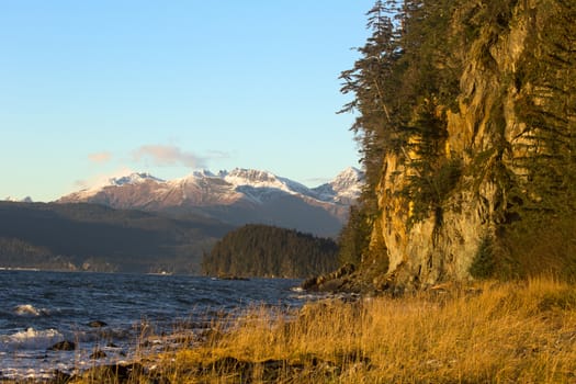 Sunlight highlights autumn colors of Fritz Cove on northwestern coast of Douglas Island, Juneau, Alaska; blue waters of Stephens Passage and mountains enhance scene of natural beauty; copy space in blue sky of horizontal image; 