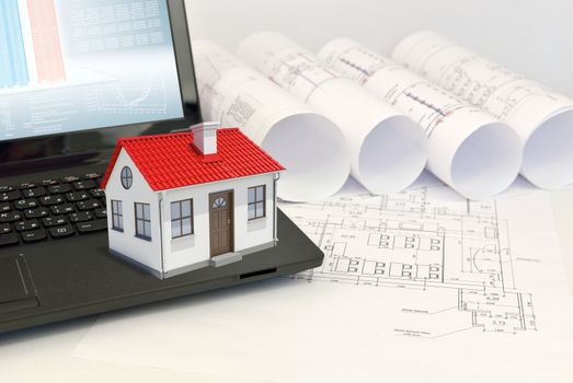 Small model house with red roof on laptop near scrolls of architectural drawings. Construction concept