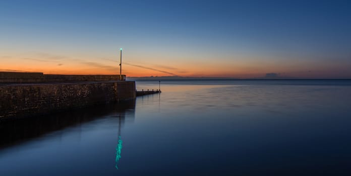 a stone breakwater, in a very calm sea, at sunset, with a green warning light, reflecting in the sea