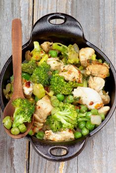 Homemade Chicken Stew with Broccoli, Bell Pepper and Green Pea in Black Saucepan with Wooden Spoon isolated on Rustic Wooden background. Top View