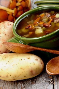 Delicious Vegetarian Soup with Chanterelle Mushrooms, Potato, Onion and Carrot in Green Pot with Raw Ingredients and Wooden Spoon closeup on Rustic Wooden background