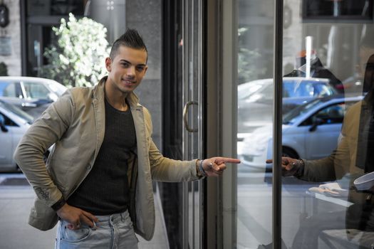 Attractive young man in urban setting, pointing finger at clothes in shop window, smiling at camera