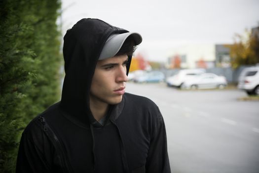 Attractive young man with hoodie and baseball cap in city street, sad or thinking
