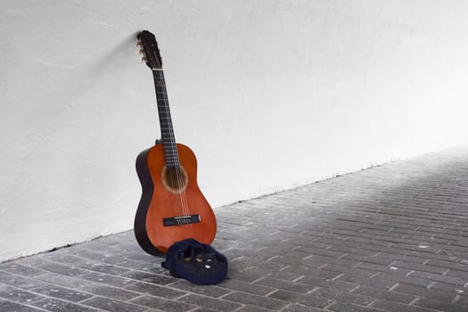 Guitar leaning against white wall with blue hat containing some coins in front 