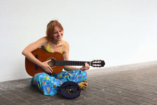 Young woman playing the guitar on the street with a hat containing some coins in front of her 