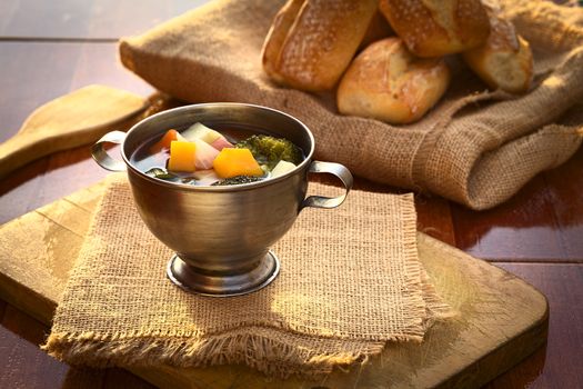 Fresh vegetarian soup made of broccoli, squash, onion, carrot, potato and tomato served in a metal cup on jute cloth and wooden board with ciabatta buns in the back, photographed with natural light (Selective Focus, Focus onto the first half of the soup)