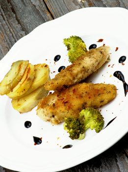 Delicious Fried Chicken Strips in Herb Breading Served with Steamed Broccoli, Roasted Potato Slices and Balsamic Sauce on White Plate closeup on Rustic Wooden background