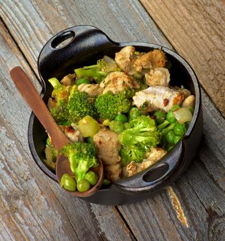 Homemade Chicken Stew with Broccoli, Bell Pepper and Green Pea in Black Saucepan with Wooden Spoon isolated on Rustic Wooden background