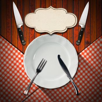 Empty and white plate on wooden brown background and red and white checkered tablecloth with silver cutlery, two kitchen knives and empty label
