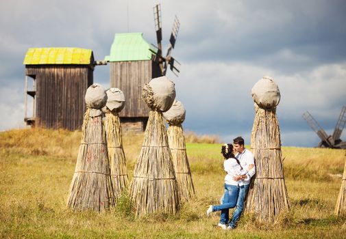 Young couple in Ukrainian style clothing kissing on field with bundles of straw and old windmills in background