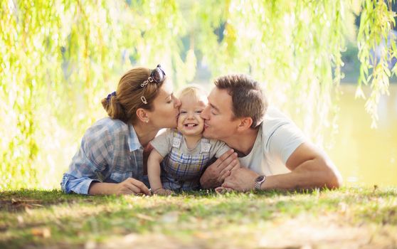 Happy parents kissing toddler boy. Father, mother and son relaxing on the lawn.