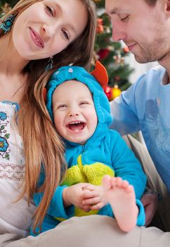 Young parents cover camera with hand while kissing, baby son dressed in little monster costume laughing, Christmas time.