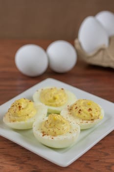 Deviled eggs on a white plate with a try of eggs in the background and sitting on a wooden table.