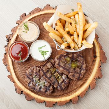 Portion of BBQ beef filet mignon steak  served  on wooden board with  ketchup, mustard and cream sauces, fried potatoes in aluminium bucket