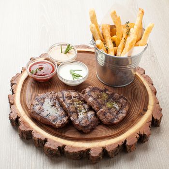 Portion of BBQ beef filet mignon steak  served  on wooden board with  ketchup, mustard and cream sauces, fried potatoes in aluminium bucket