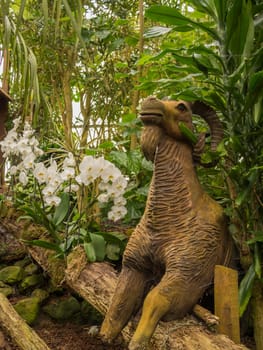 Statue of a wooden ram in a forest besides some blooming orchids