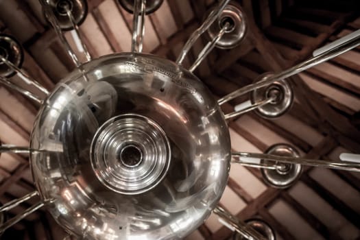 A close up black and white photo of a chandelier in a church with a beamed ceiling in the background.