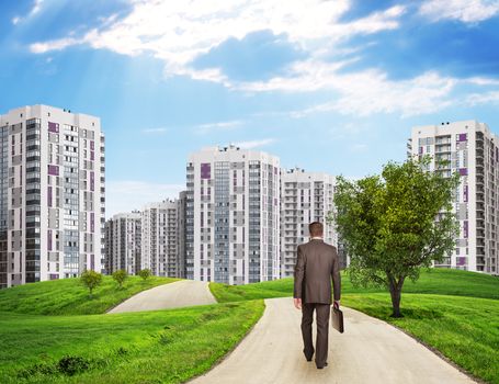 Businessman walking along road running through green hills with a few trees. High-rise buildings as backdrop 