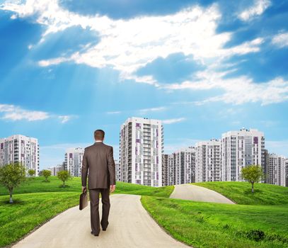 Businessman walking along road running through green hills with a few trees. High-rise buildings as backdrop 