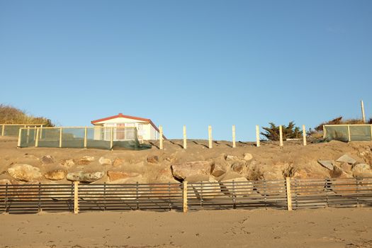 An area of constructed sand dune with a sand drift fence on wooden posts in front of a row of large rocks banked up with sand, a fence and holiday home on top.