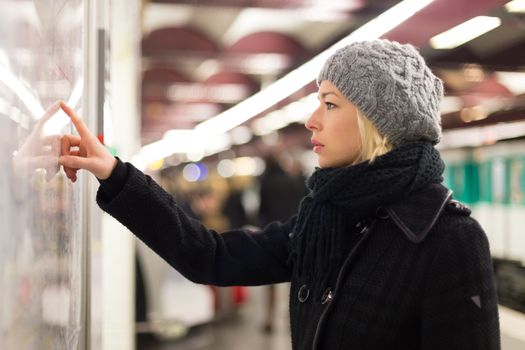 Casually dressed woman wearing winter coat, orientating herself with public transport map panel, pointing on her final destination. Urban transport.