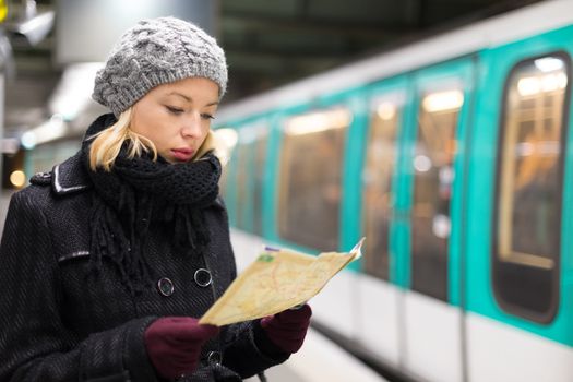 Casually dressed woman wearing winter coat, waiting on a platform for a train to arrive, orientating herself with public transport map. Urban transport.