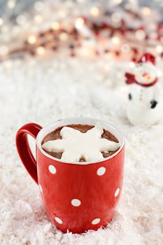 Vibrant red cup of hot chocolate with snow flake shape of whipped cream. Snowman and Christmas lights in the background. Extreme shallow depth of field.