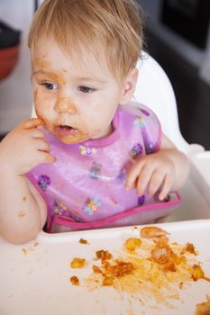 blonde caucasian baby 1 year eating tomato meal with her hand in high-chair