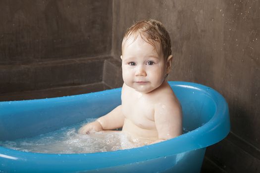 blonde naked baby washing in blue little bath indoor with brown background