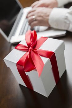 White Gift Box with Red Ribbon and Bow Near Man Typing on Laptop Computer.