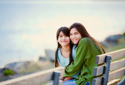 Two happy biracial young girls sitting together on bench next to lake