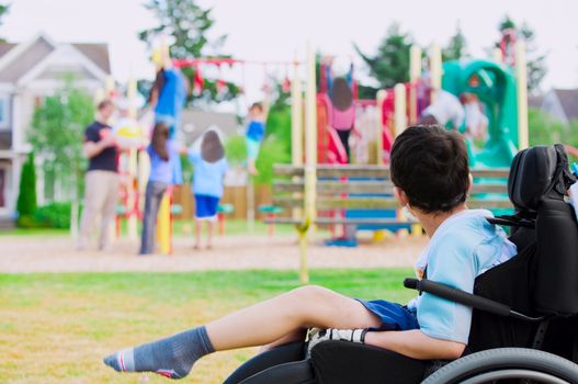 Disabled little boy in wheelchair sadly watching children play on playground