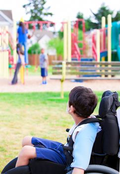 Disabled little boy in wheelchair sadly watching children play on playground