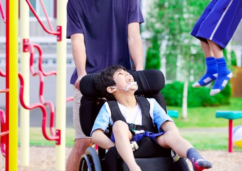 Disabled boy in wheelchair enjoying watching friends play at park on jungle gym