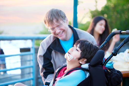 Father talking with disabled son in wheelchair outdoors by lake