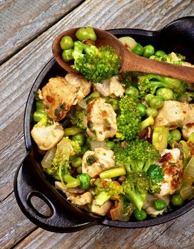 Homemade Chicken Stew with Broccoli, Bell Pepper and Green Pea in Black Saucepan with Wooden Spoon closeup on Rustic Wooden background. Top View