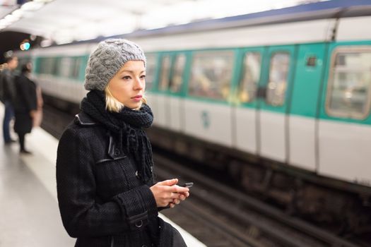 Young woman in winter coat with a cell phone in her hand waiting on the platform of a railway station for train to arrive. Public transport.  