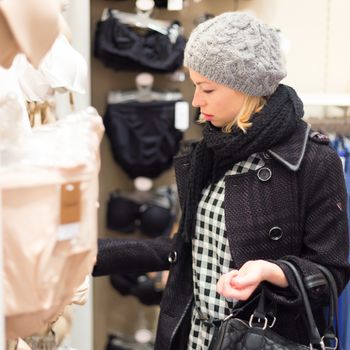 Woman shopping lingerie . Shopper looking and choosing clothing indoors in store. Beautiful blonde caucasian female model wearing winter coat and fashionable knitted cap.