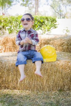 Cute Young Mixed Race Boy Laughing with Sunglasses and Hard Hat Outside Sitting on Hay Bale.