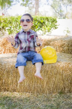Cute Young Mixed Race Boy Laughing with Sunglasses and Hard Hat Outside Sitting on Hay Bale.