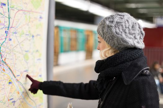 Casually dressed woman wearing winter coat, orientating herself with public transport map panel, pointing on her final destination. Urban transport.