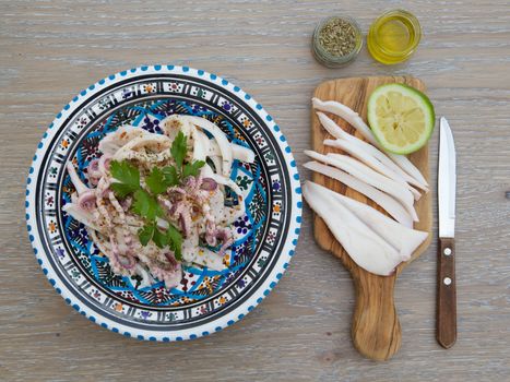 Marinated squid salad with fresh parsley in the traditional Tunisian plate. Oregano, olive oil and squeezed lemon, cut peaces of squid on the wooden cutting board and a knife close to the plate on the wooden surface.