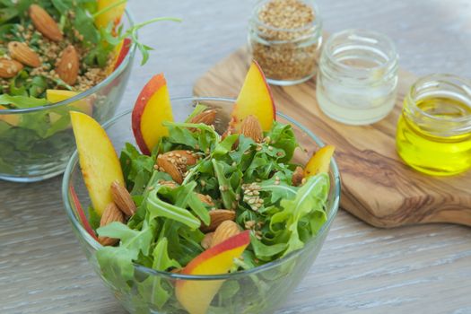 Vitamin salad- rucola with almond, peach and sesame seeds in glass dish. Olive oil, lemon juice and sesame seeds on the wooden cutting board in the background. Close up