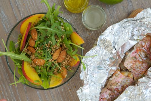 Vitamin salad- rucola with almond, peach and sesame seeds in glass dish and baked fish in the aluminium foil from the right side. Top view