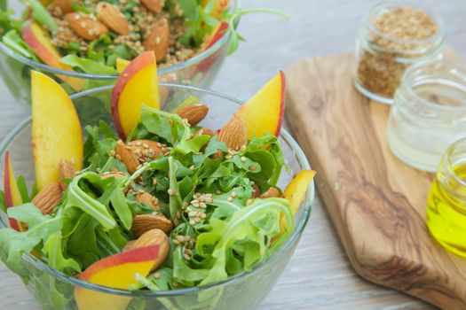 Vitamin salad- rucola with almond, peach and sesame seeds in glass dish. Olive oil, lemon juice and sesame seeds on the wooden cutting board in the background. Close up